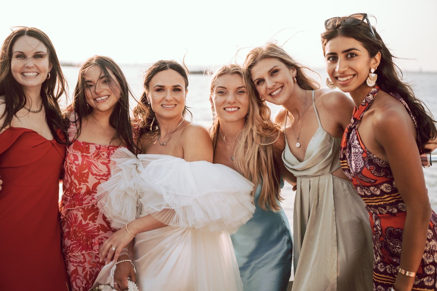 Six women in elegant dresses pose together smiling at an outdoor wedding celebration near the water.