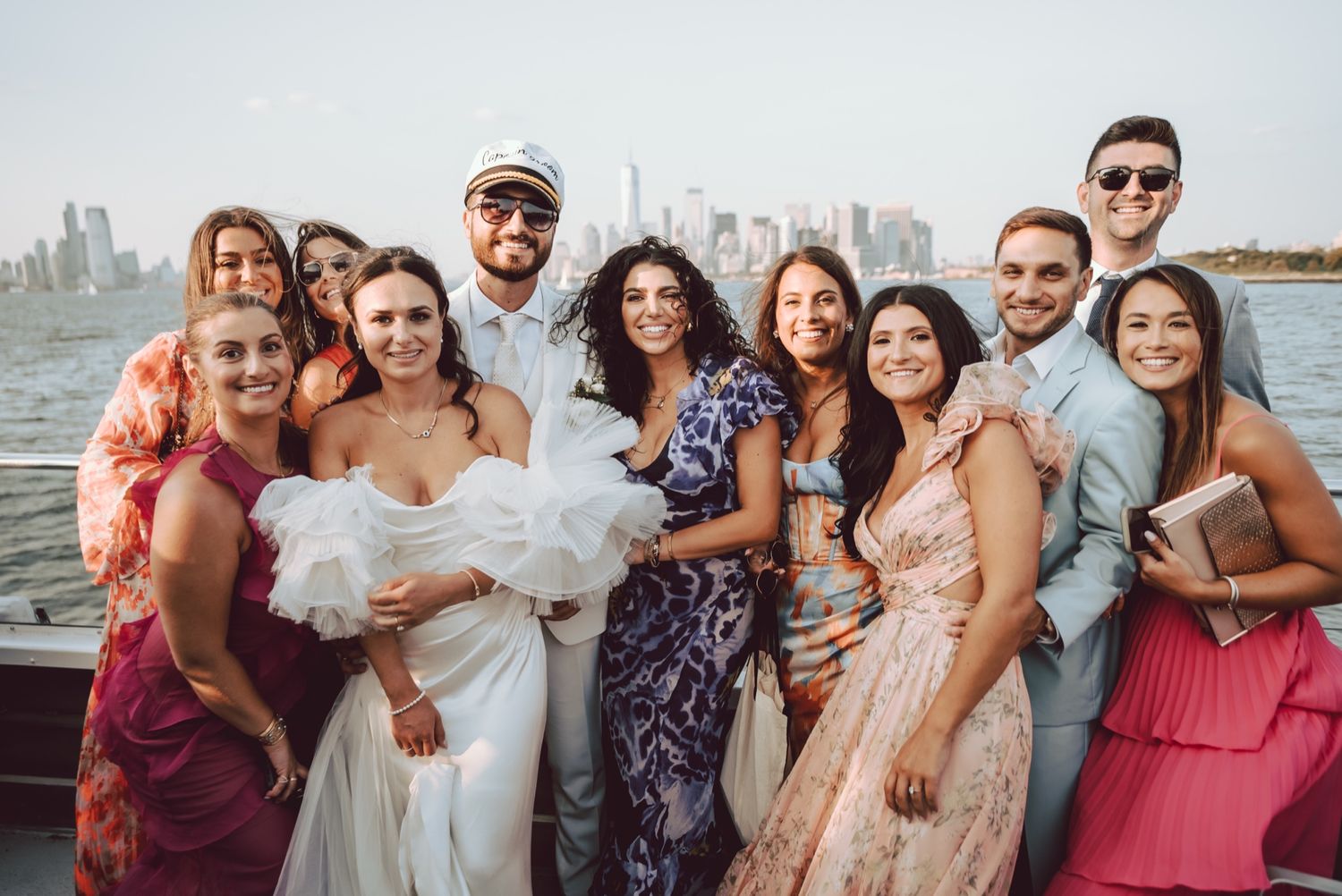 Wedding party poses on a boat with NYC skyline backdrop, bride in white gown surrounded by smiling guests.