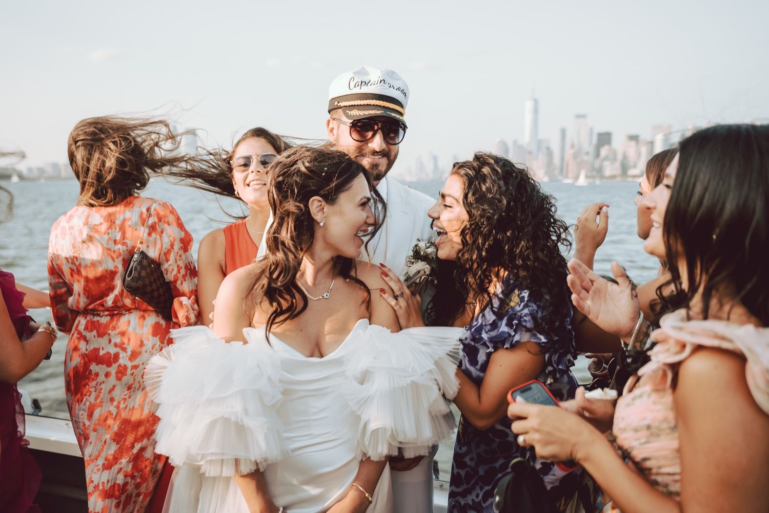 Bride in white ruffled dress laughing with guests on a boat wedding celebration with NYC skyline in background.