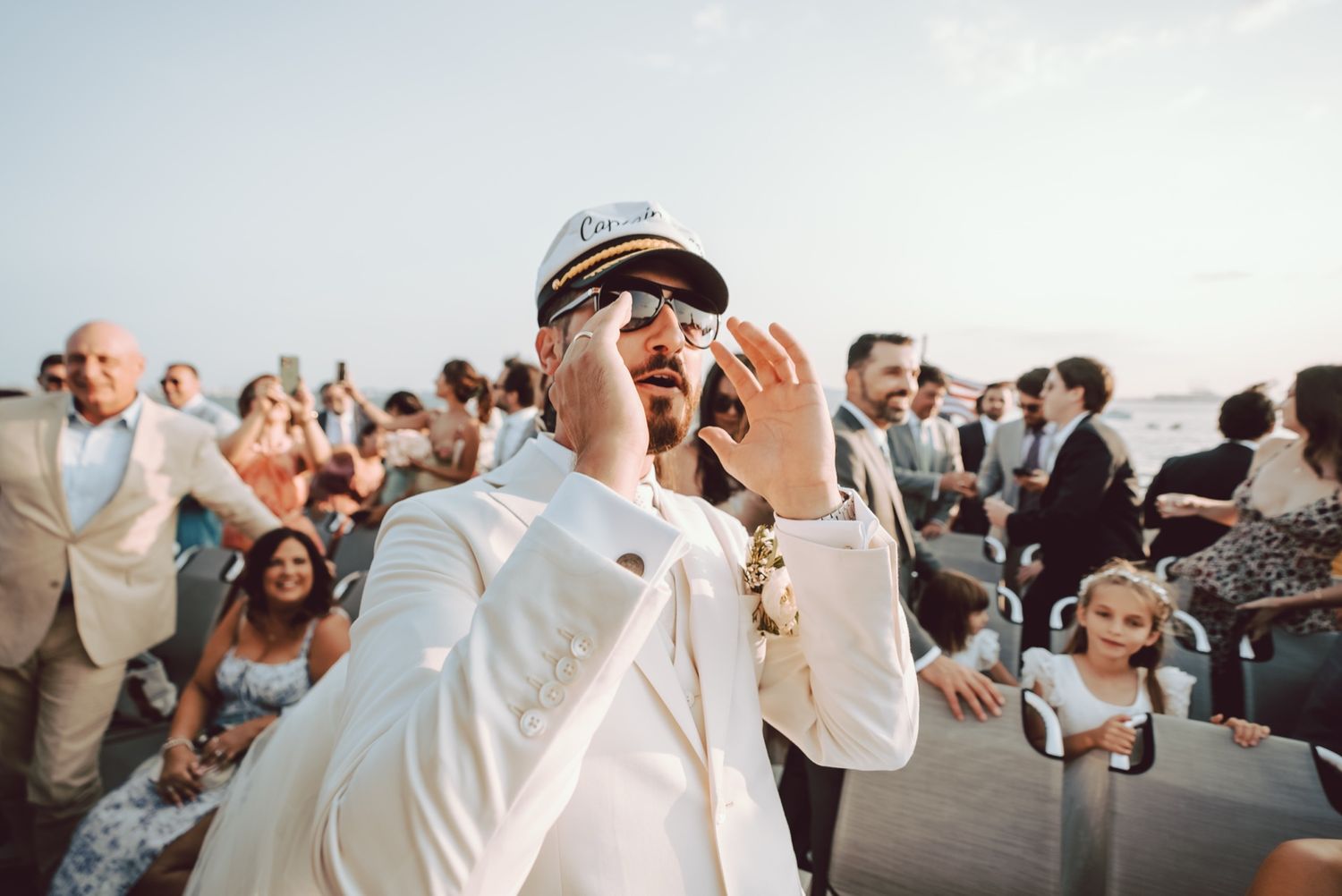 Groom in white suit and captain hat adjusts sunglasses at outdoor wedding celebration with guests.