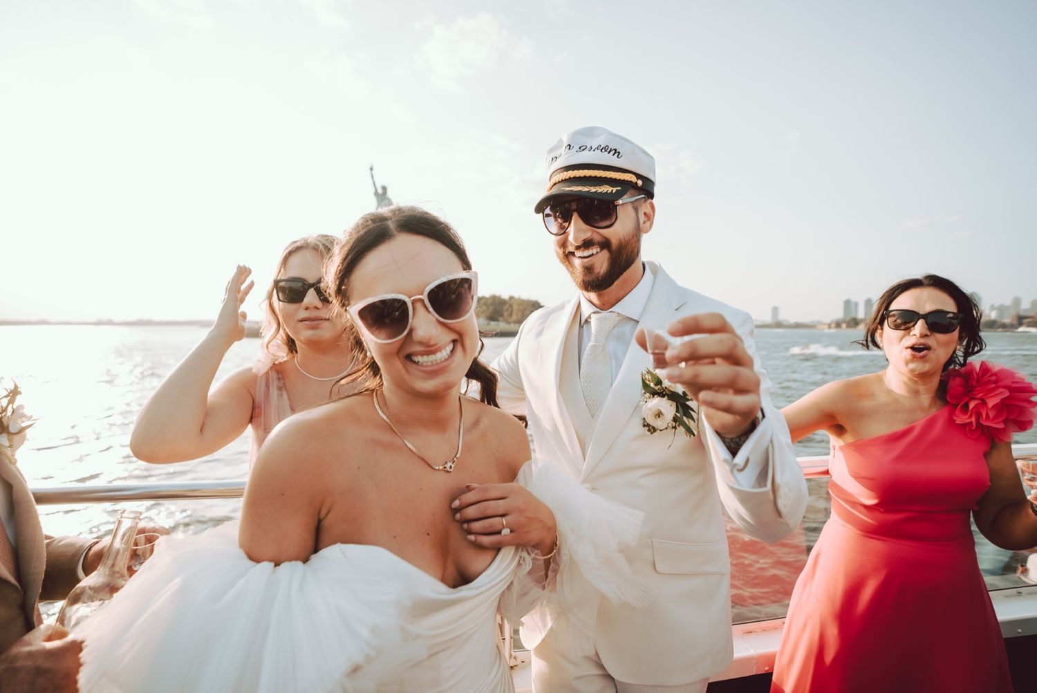 Bride and groom in sunglasses and captain hat celebrating on a boat with guests.