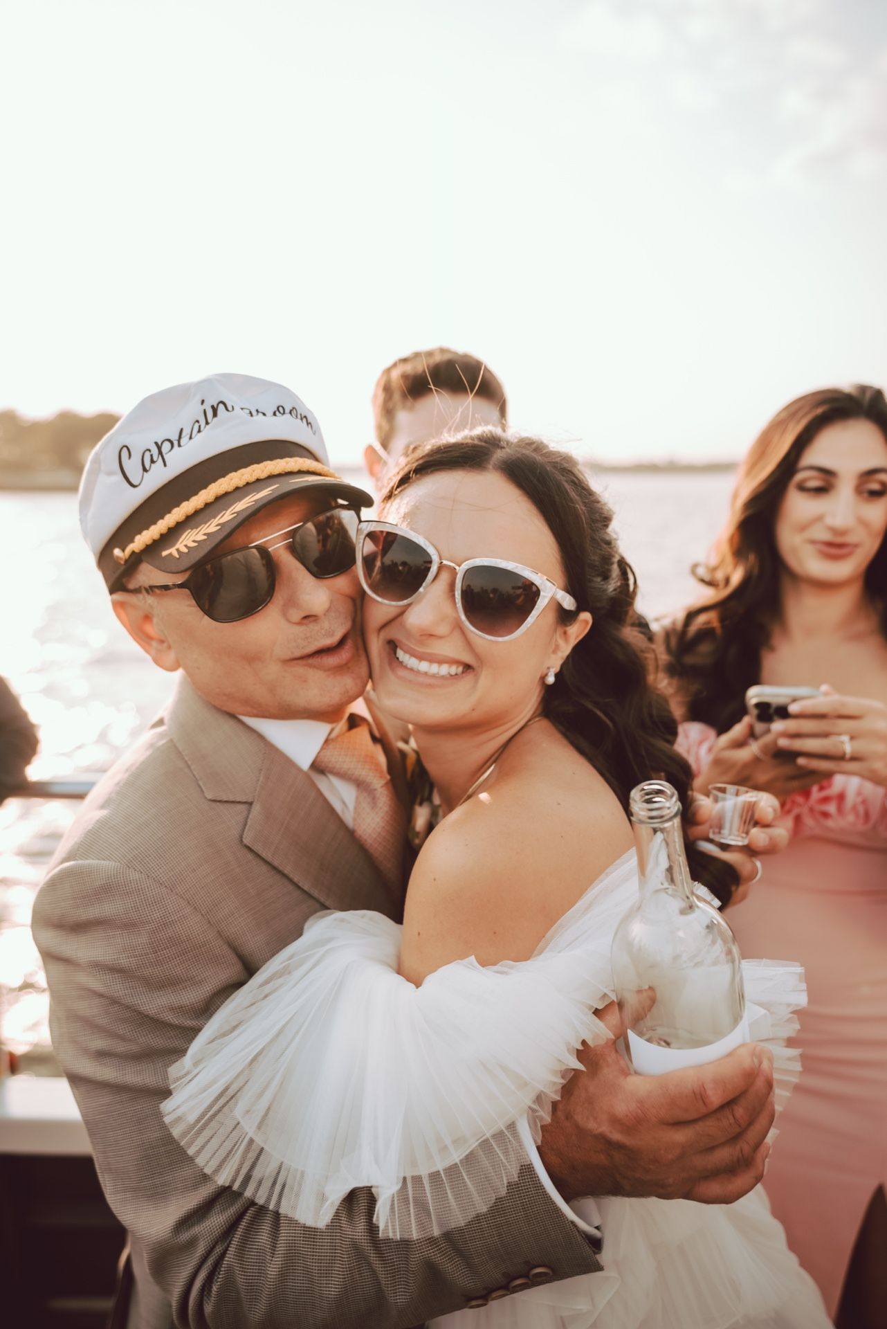 Groom in captain hat hugging bride holding champagne bottle on a sunny boat wedding.