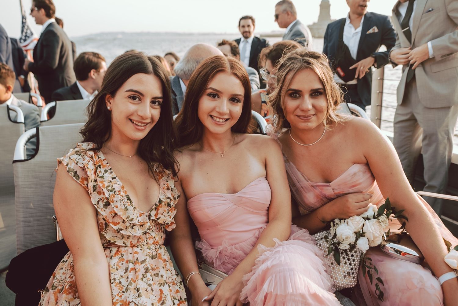 Three women in pink dresses smile together at an outdoor wedding celebration.