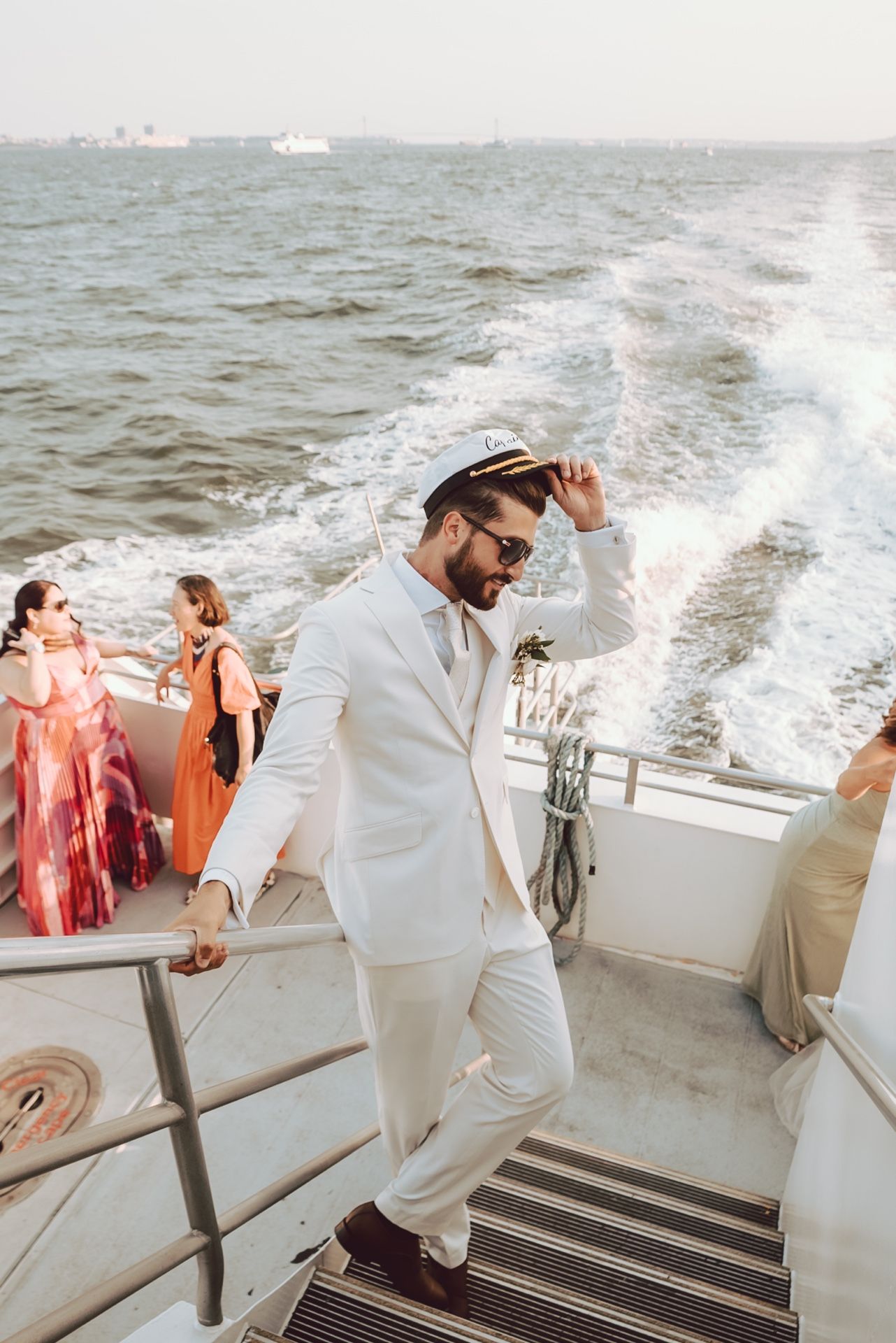 Groom in white suit and captain hat posing on the deck of a moving boat.