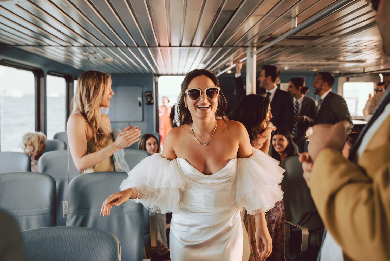 Bride in off-shoulder wedding dress walking through boat interior wearing sunglasses.