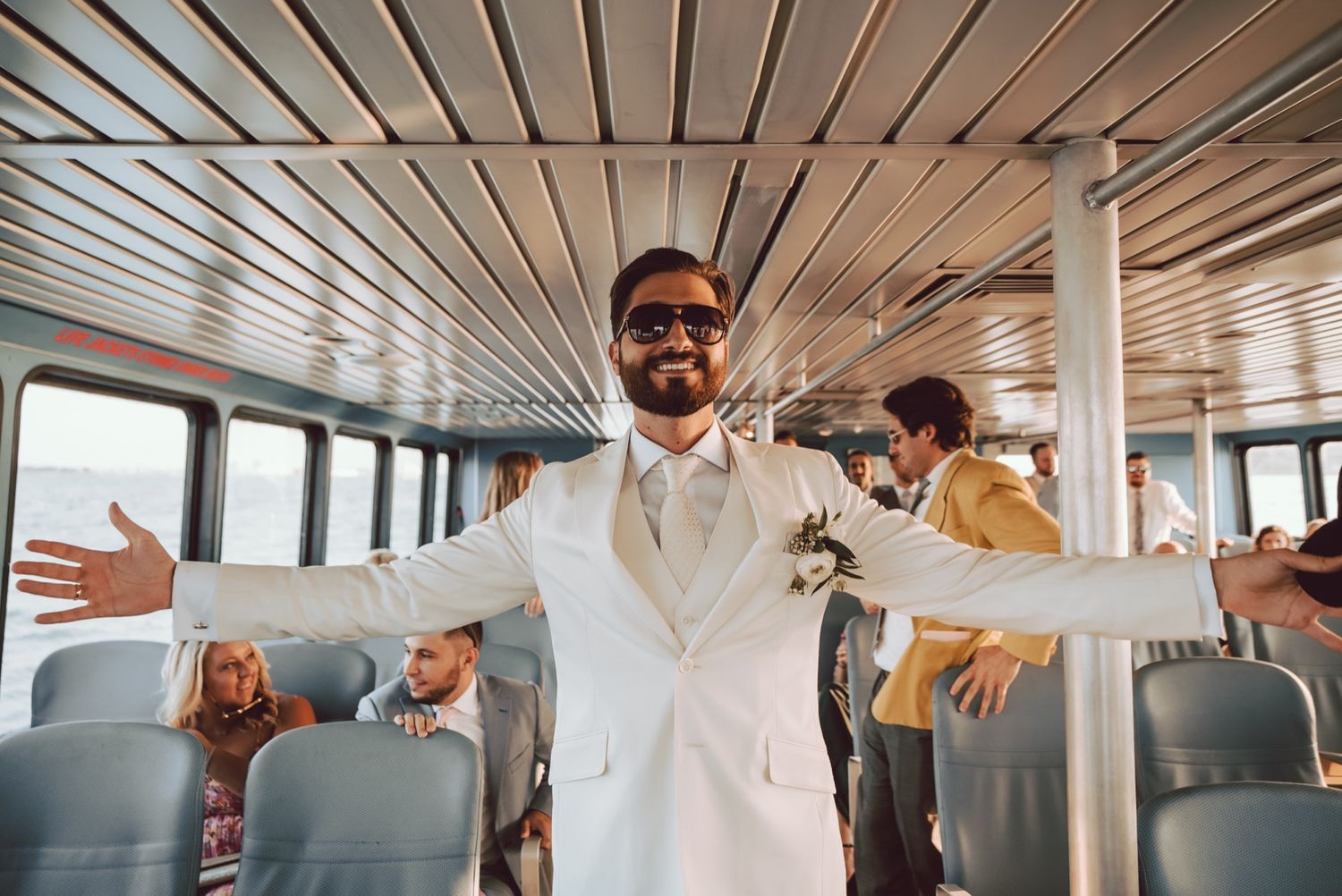 Groom in white suit spreading arms wide open inside a boat cabin at wedding.