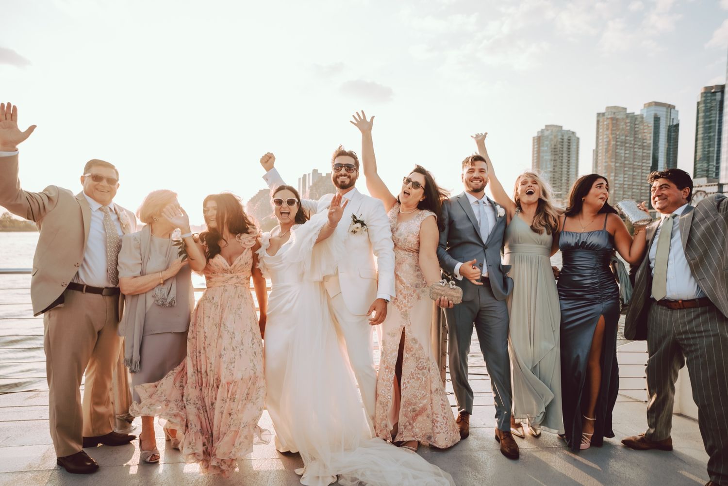Wedding party celebrating with raised arms on a waterfront dock with city skyline.