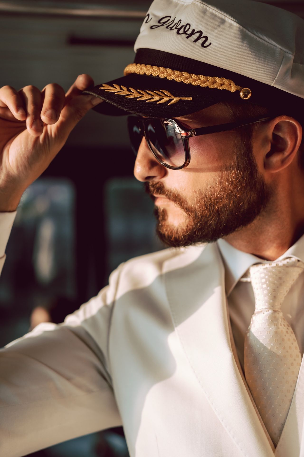 A stylish bearded man in a white suit and captain's hat adjusts his sunglasses with a confident expression.