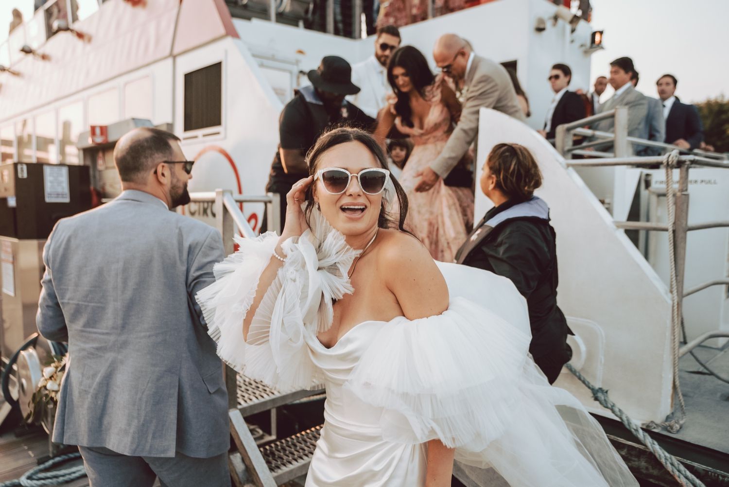 Bride in white ruffled off-shoulder gown with sunglasses boarding a wedding boat.