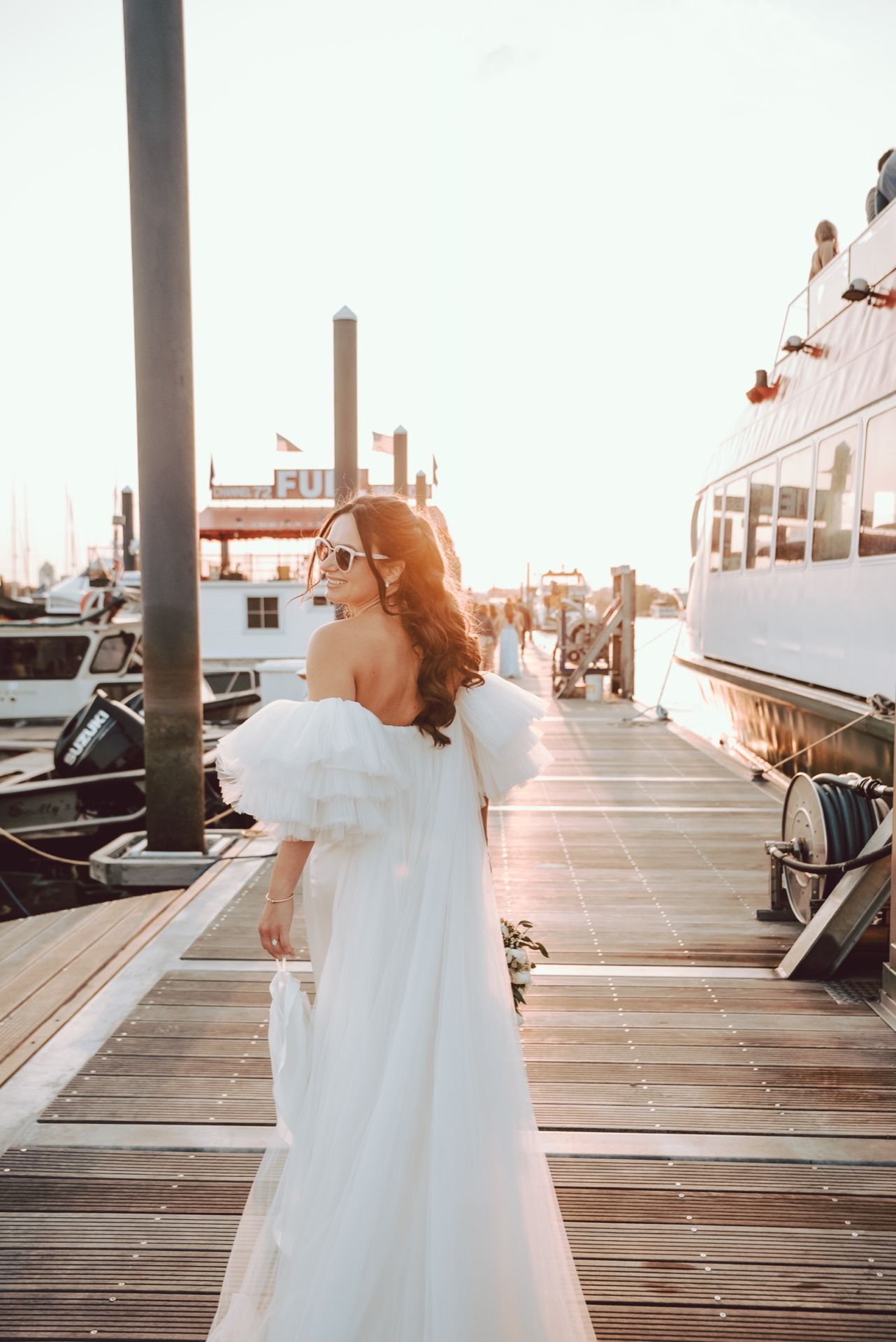 Bride in elegant white ruffled gown posing on sunny marina dock beside luxury yacht.