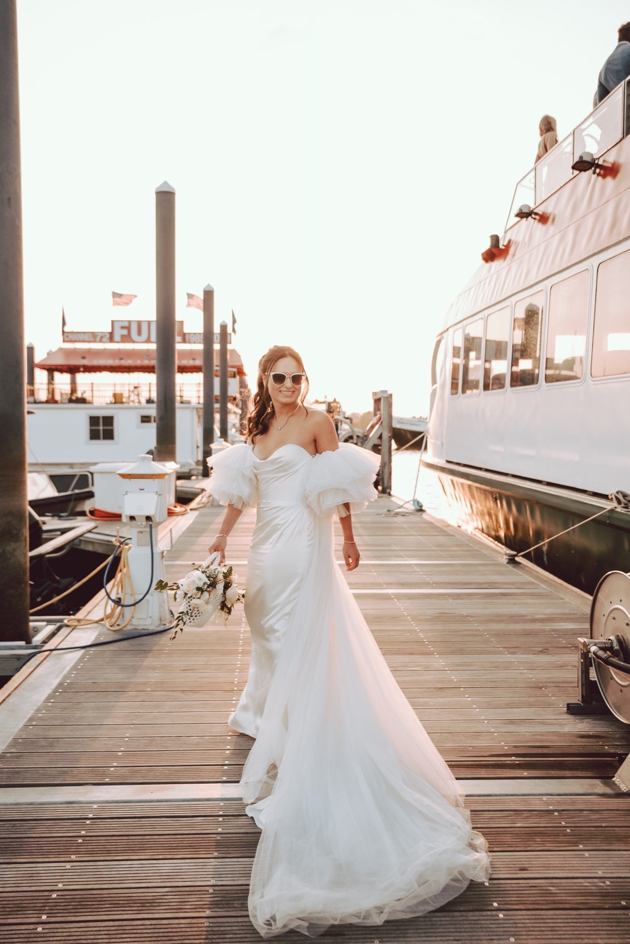 Bride in white off-shoulder gown with feather sleeves walks on marina dock holding bouquet, yachts in background.