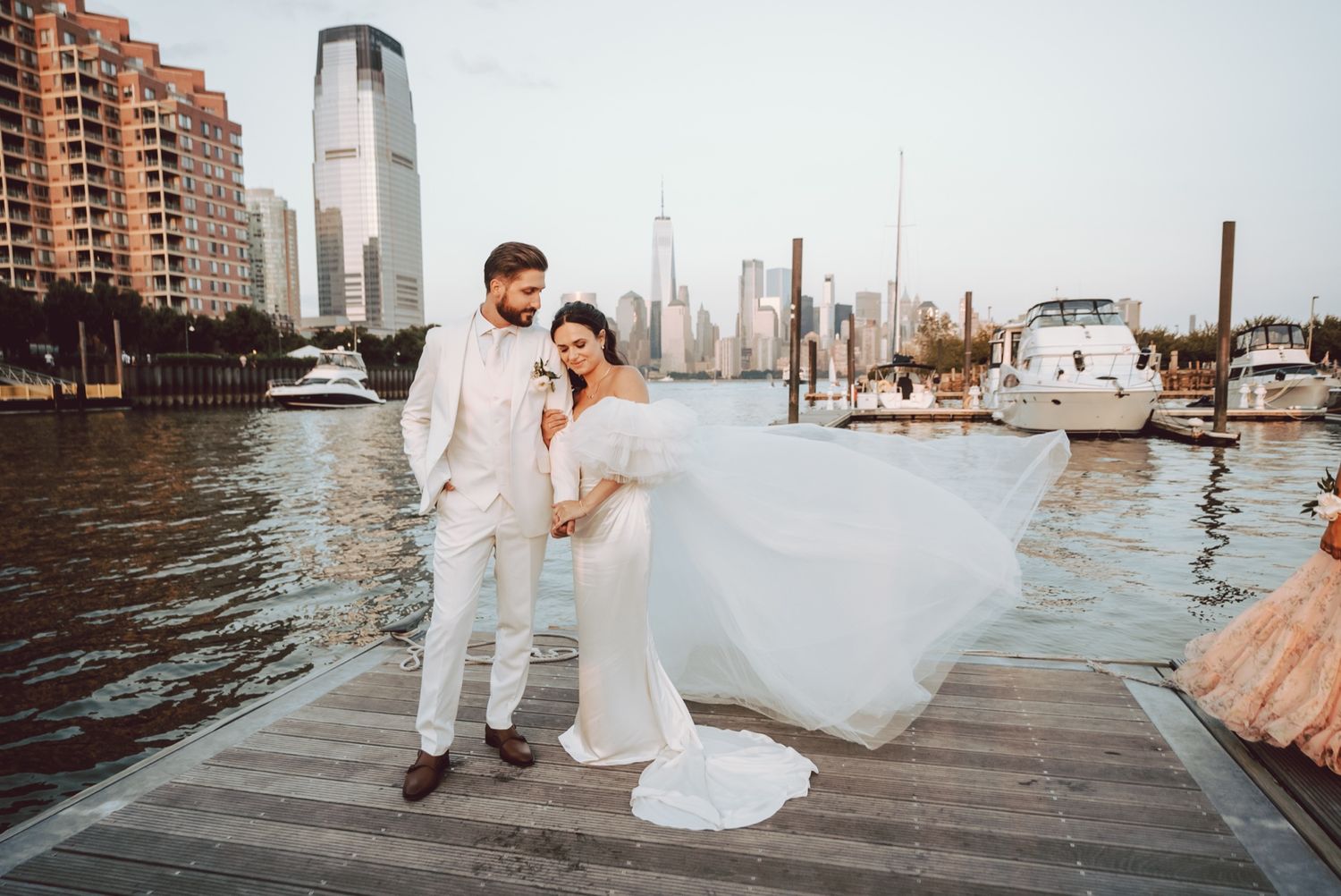 Bride and groom pose on a waterfront dock with NYC skyline backdrop, bride's veil flowing in the wind.