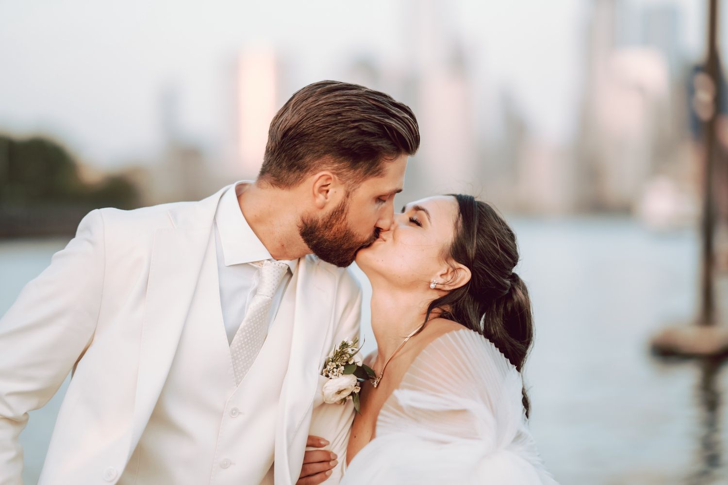 Bride and groom sharing a romantic kiss in white wedding attire with a city skyline in the background.