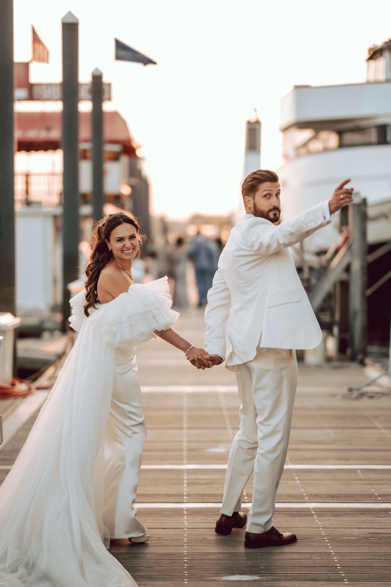 Bride and groom in white attire hold hands on a marina dock at sunset, groom pointing forward.