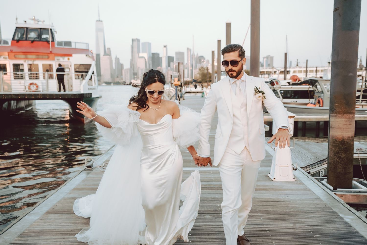 Bride and groom walking hand-in-hand on a dock with NYC skyline backdrop, both wearing sunglasses on their wedding day.