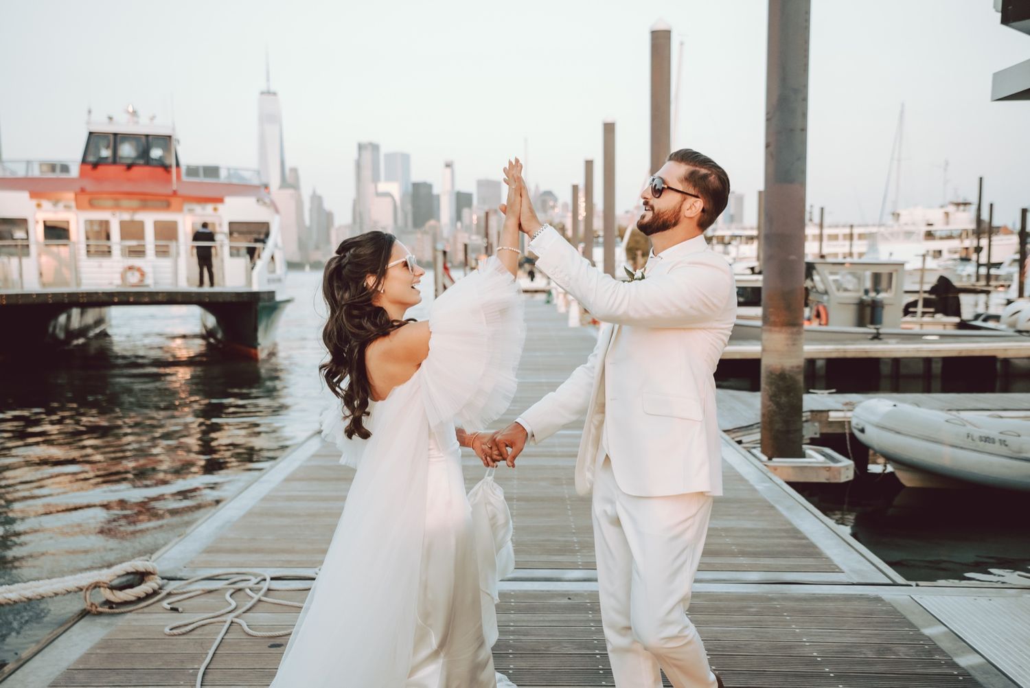 Bride and groom dancing joyfully on a waterfront dock with a marina and city skyline behind them.