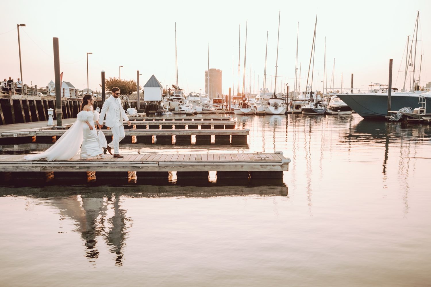 A bride and groom pose on a marina dock at golden hour, with sailboats reflected in calm water behind them.