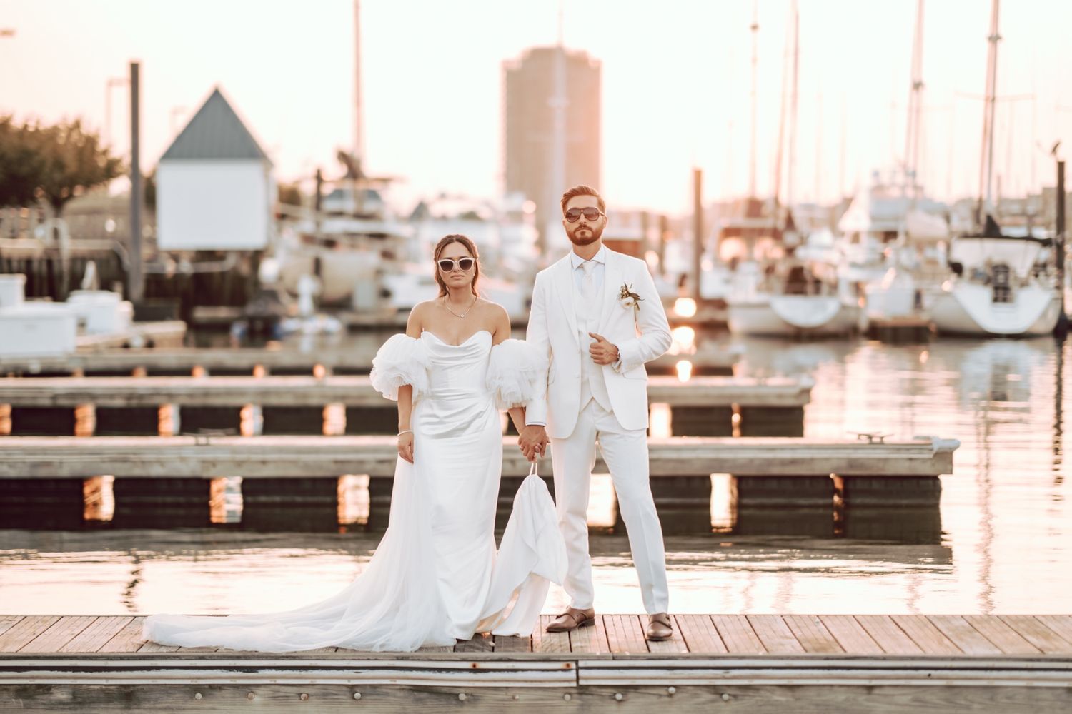 Bride and groom pose in matching white outfits with sunglasses on a marina dock at golden hour.