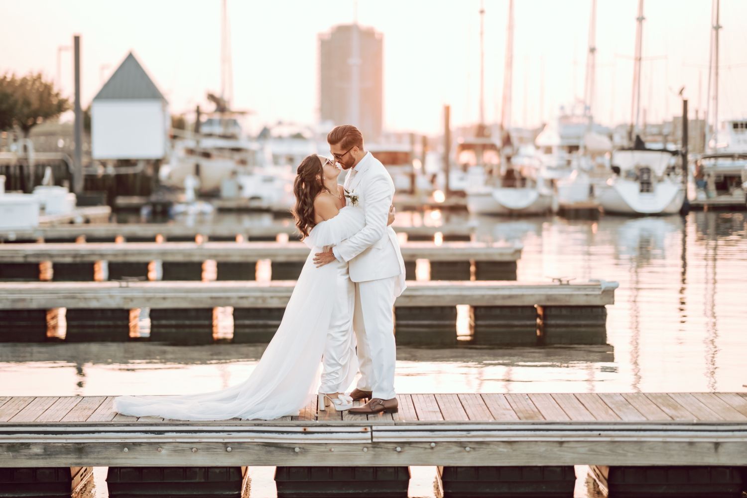 A bride and groom share a romantic kiss on a wooden dock at sunset with sailboats and a marina in the background.