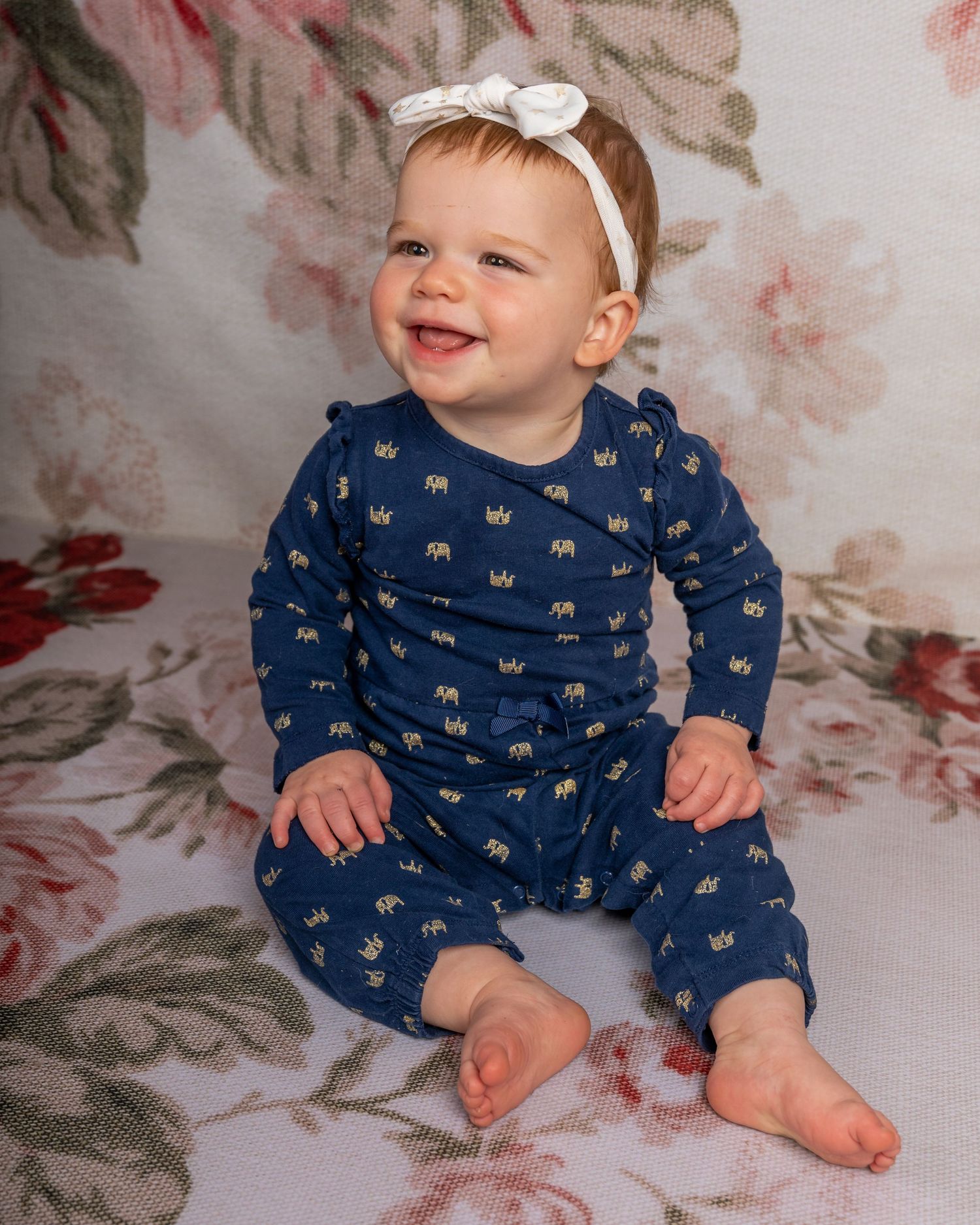 Smiling baby girl in navy blue elephant print romper with white bow headband sitting on floral background.