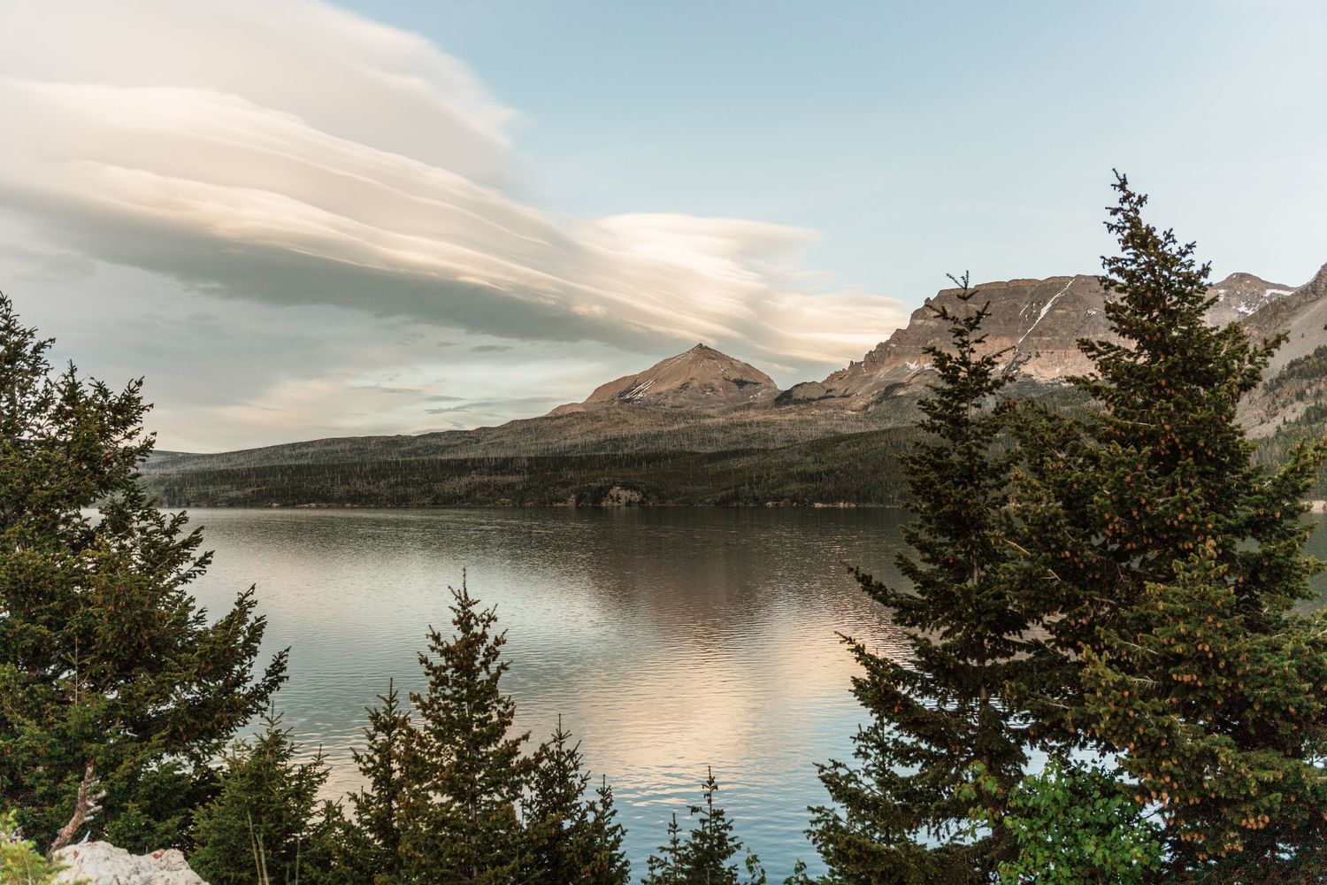 Serene mountain lake reflecting pine trees and peaks under a dramatic sky with lenticular clouds at golden hour.