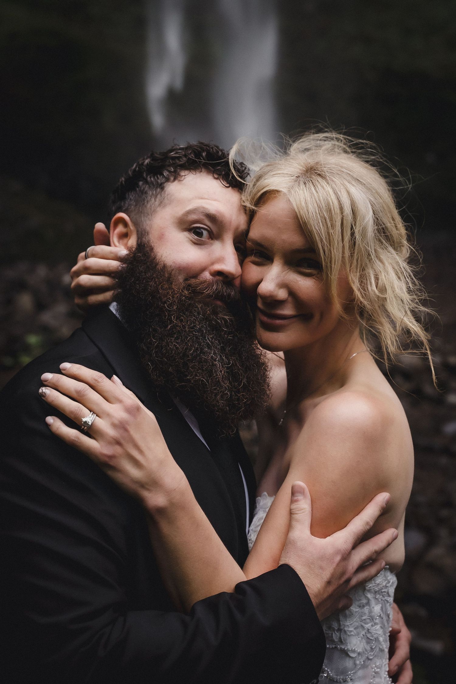 Bride and groom embrace and smile in front of a waterfall, wearing wedding attire in a lush outdoor setting.