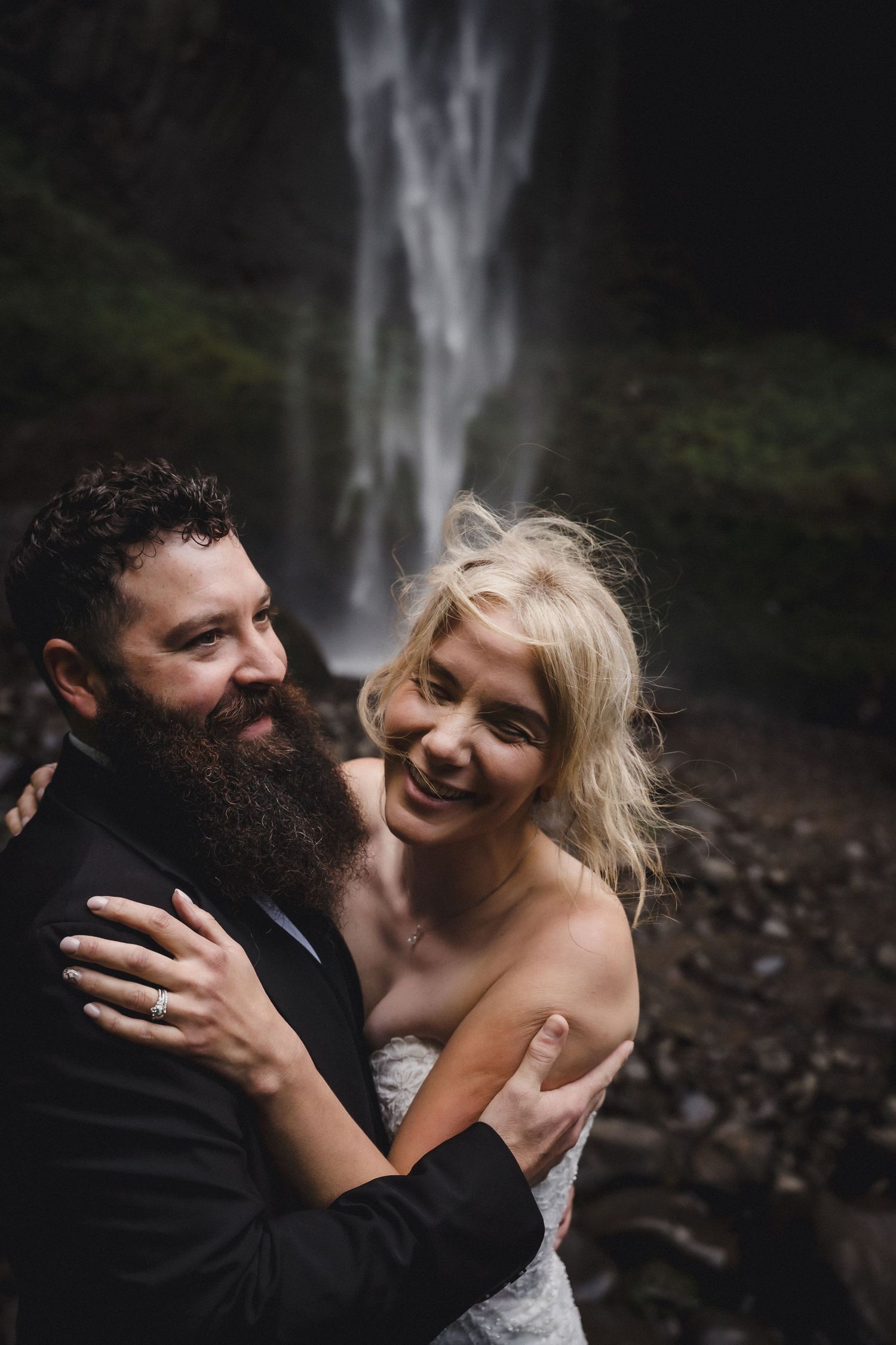 A smiling bride and bearded groom embrace in front of a waterfall, dressed in wedding attire.