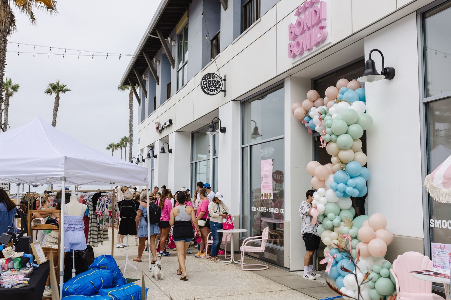 Bondi Bowls storefront grand opening with pastel balloon arch and customers gathered outside on a sunny day.