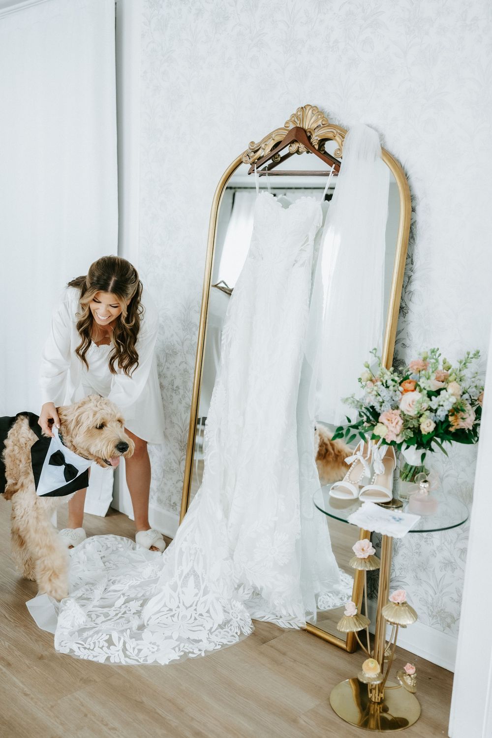Bride in white robe with dog beside elegant gold mirror displaying lace wedding gown and floral arrangement.
