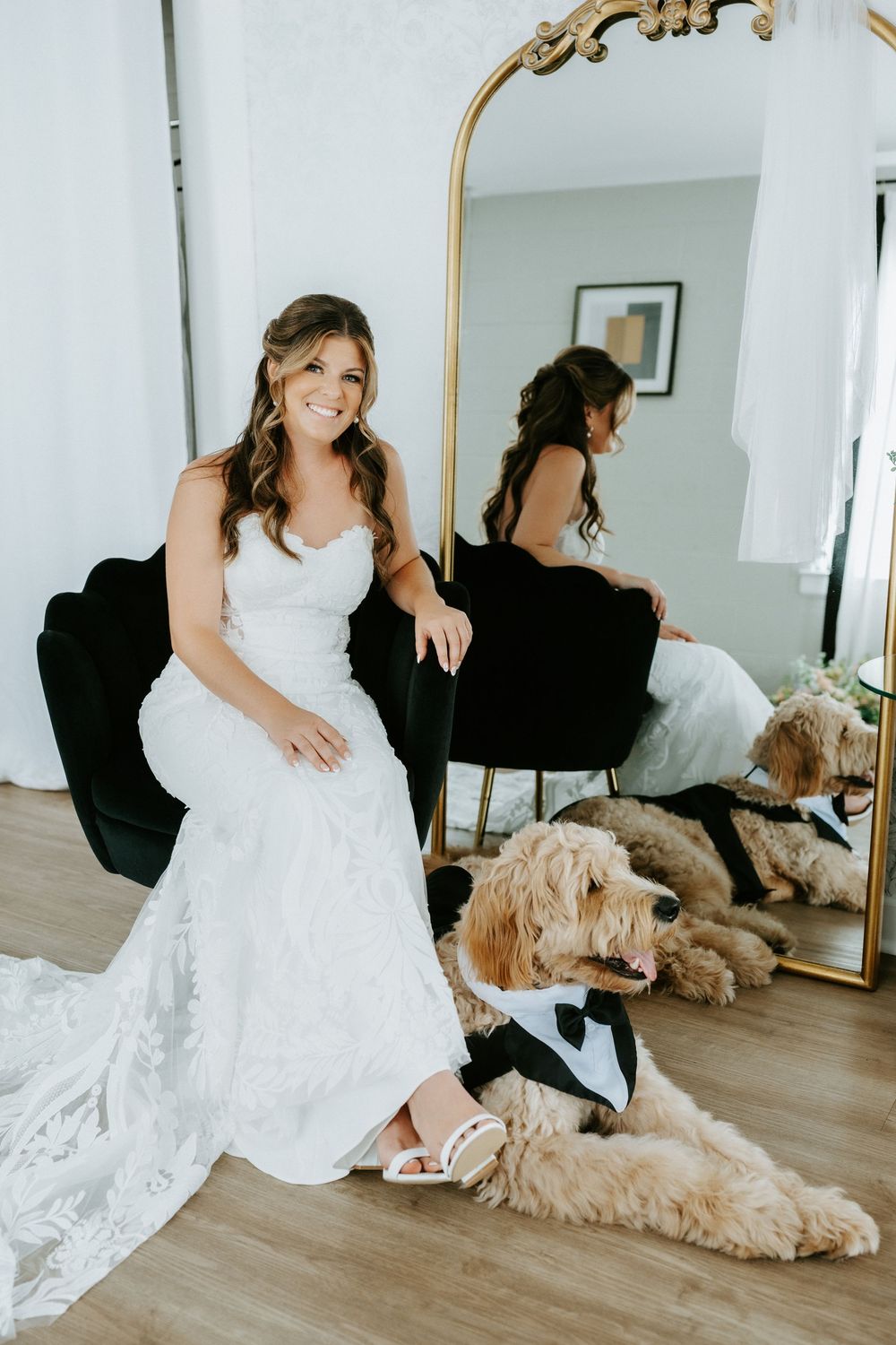 Smiling bride in white lace gown poses with golden doodle in tuxedo outfit beside ornate gold mirror.