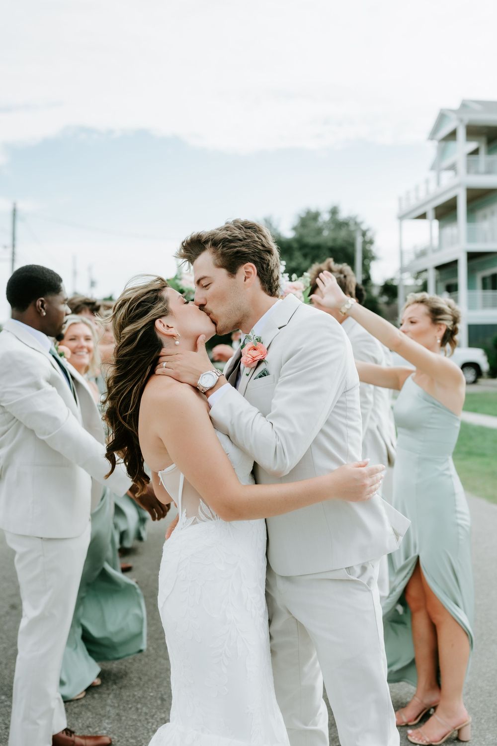 Bride and groom share a romantic kiss surrounded by wedding party in sage green dresses and white suits outdoors.