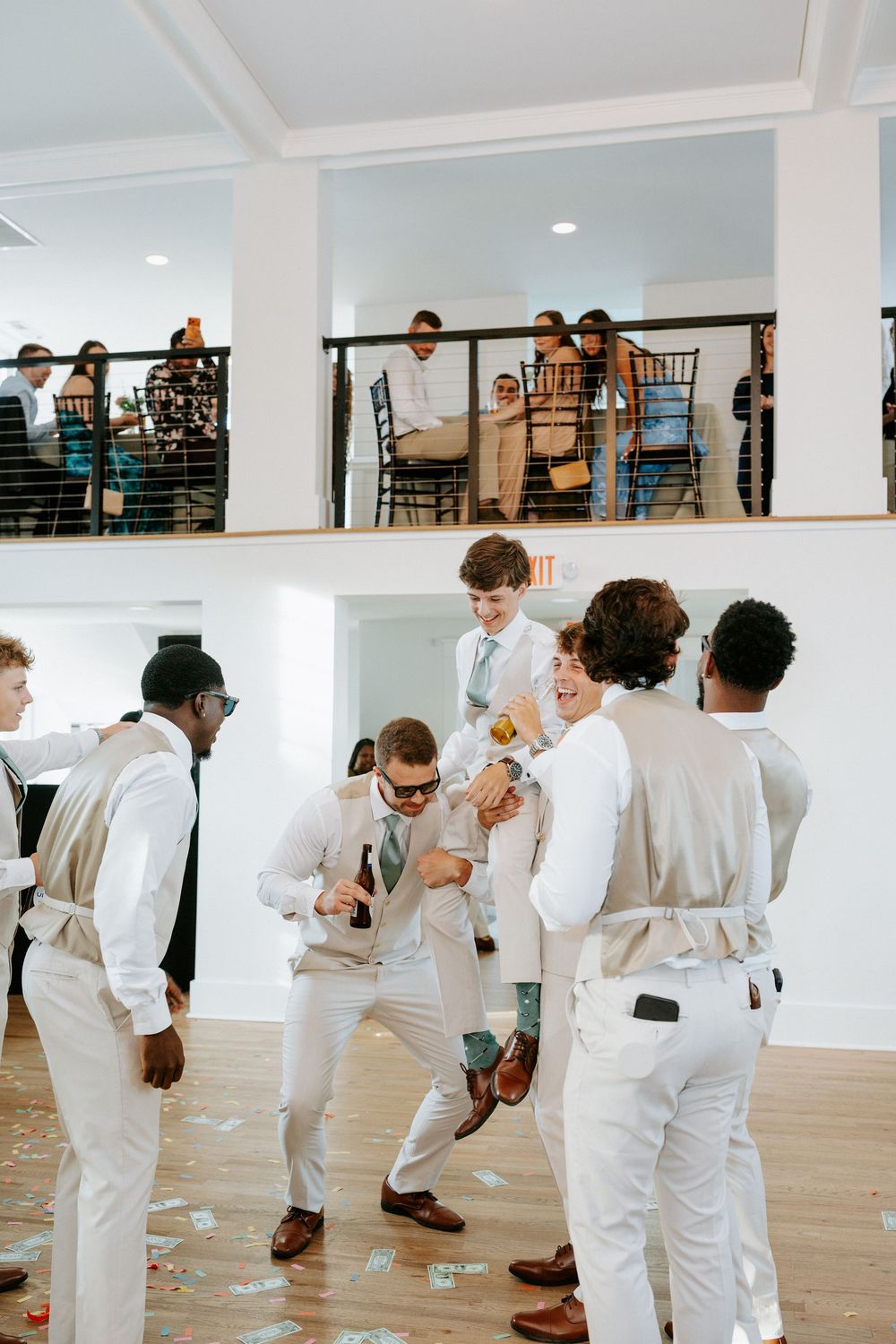Groomsmen in white suits dancing joyfully at a wedding reception with guests watching from a balcony above.