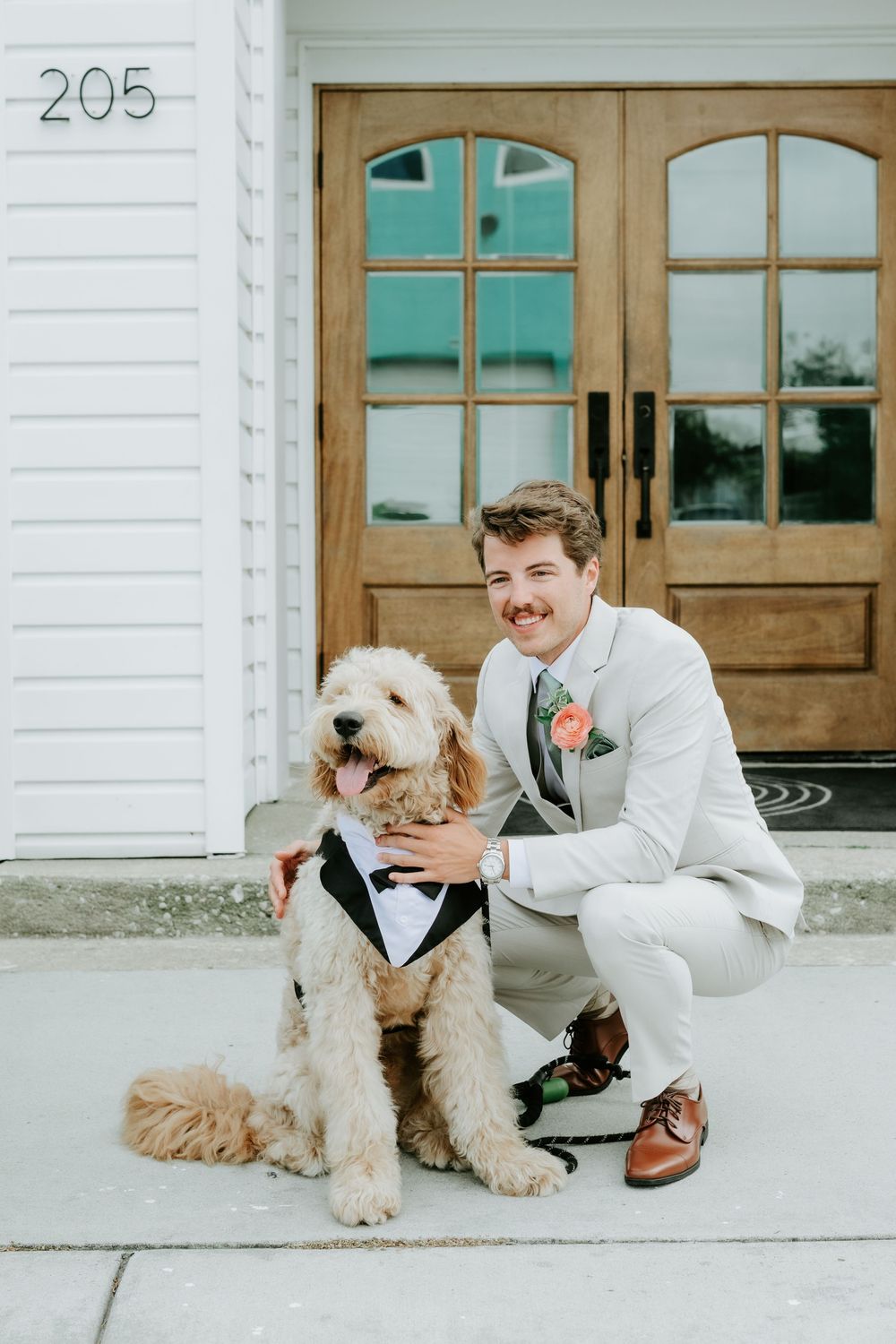 Smiling groom in white suit poses with golden doodle dog wearing tuxedo bandana outside wooden doors at address 205.