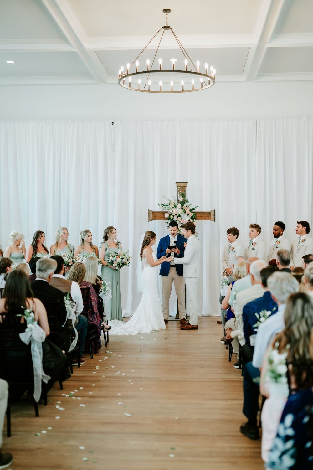 Elegant indoor wedding ceremony with couple at floral cross altar, bridesmaids, groomsmen, and guests seated in white-draped venue.