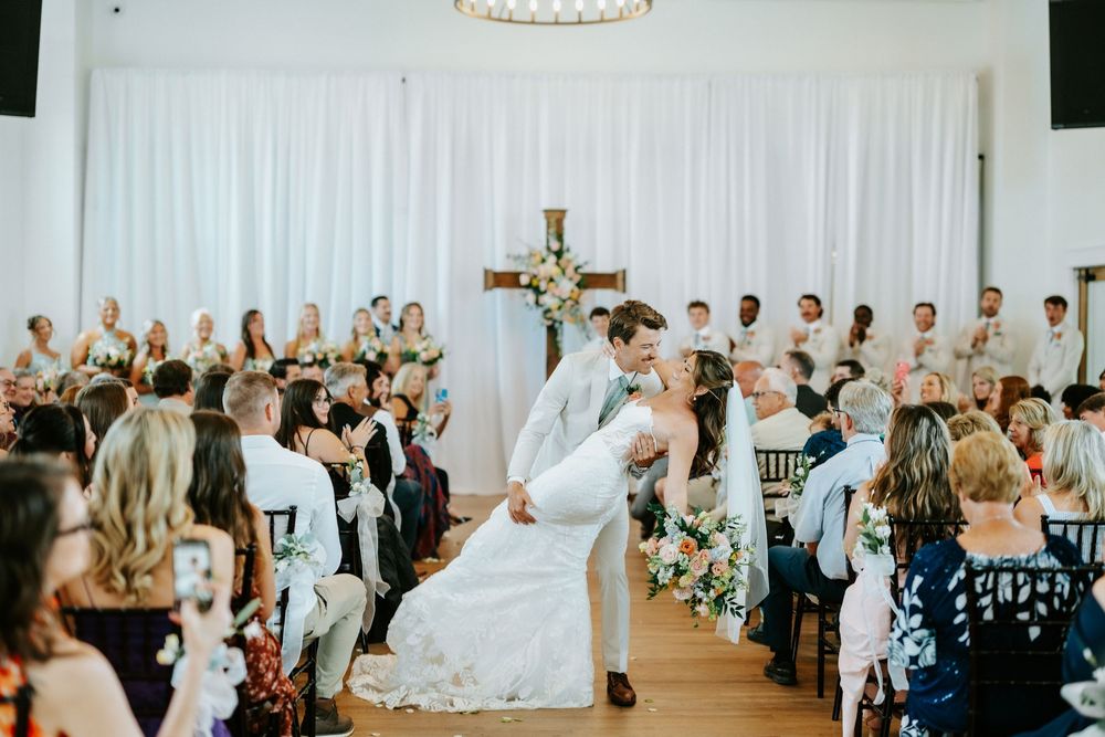 Bride and groom share a romantic dip kiss at elegant indoor wedding ceremony with floral cross and seated guests.