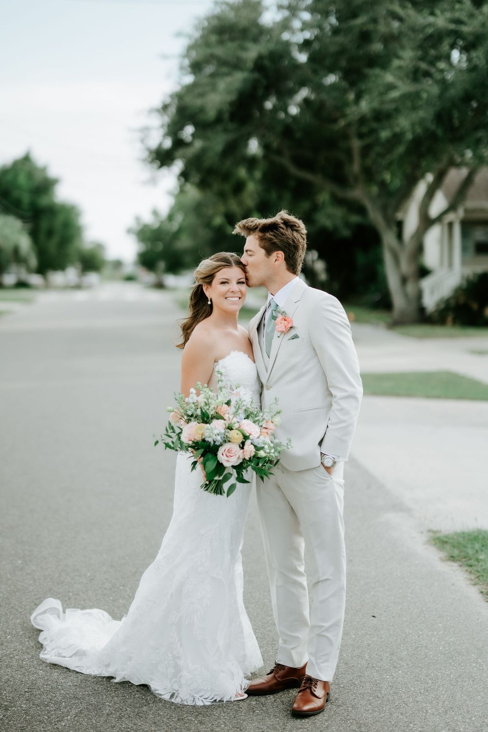 Bride in white lace gown holds pink bouquet as groom in cream suit kisses her cheek on a tree-lined street.