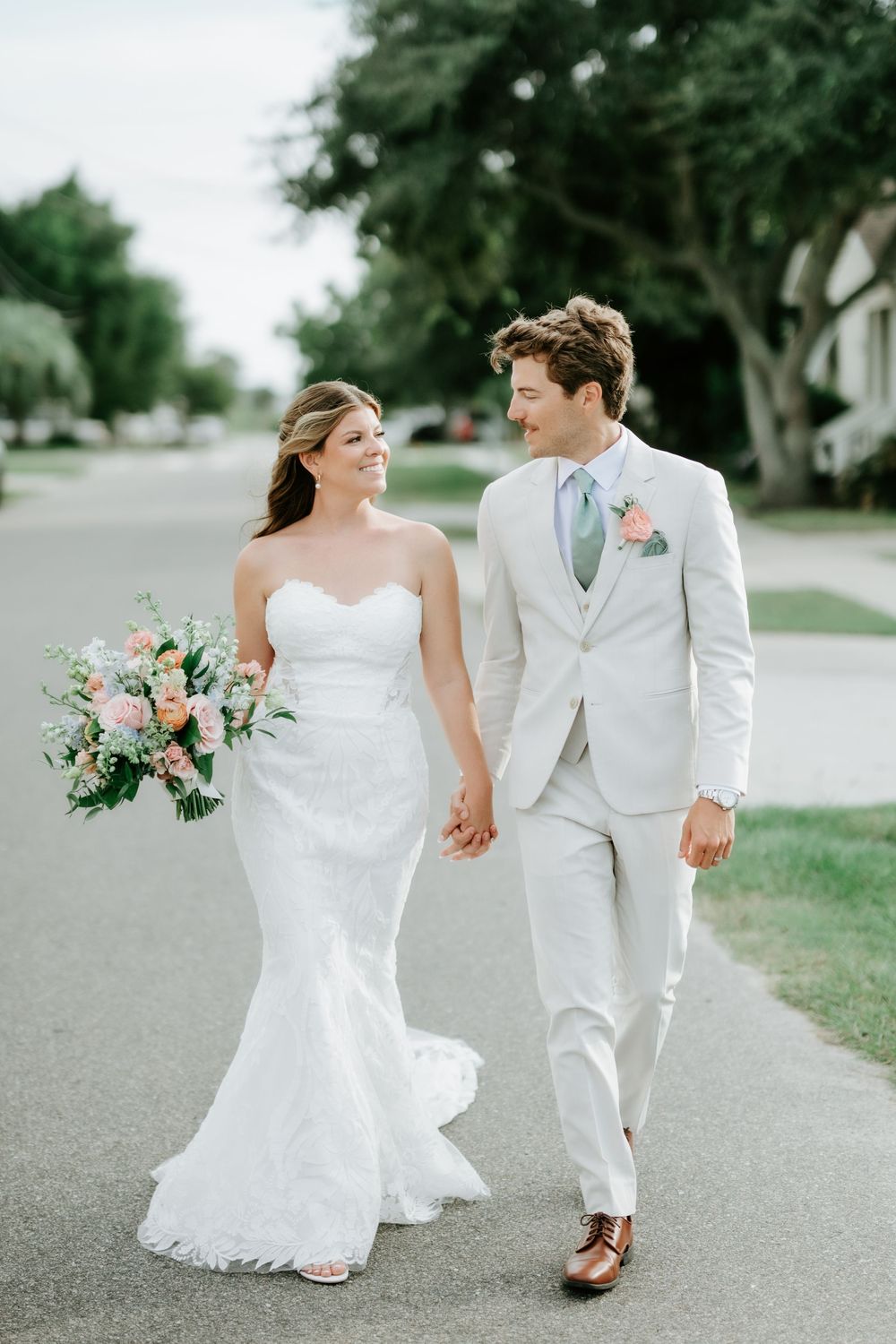Bride and groom walk hand-in-hand outdoors, she in a strapless mermaid gown, he in a light beige suit.