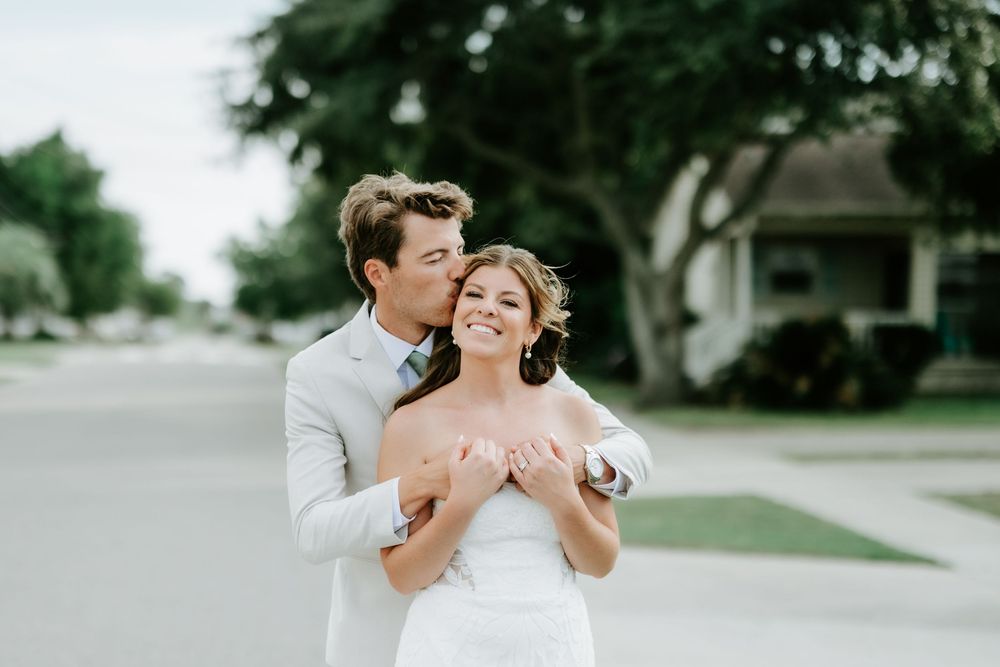 A groom in a light gray suit kisses his bride in a white strapless gown on a tree-lined street.