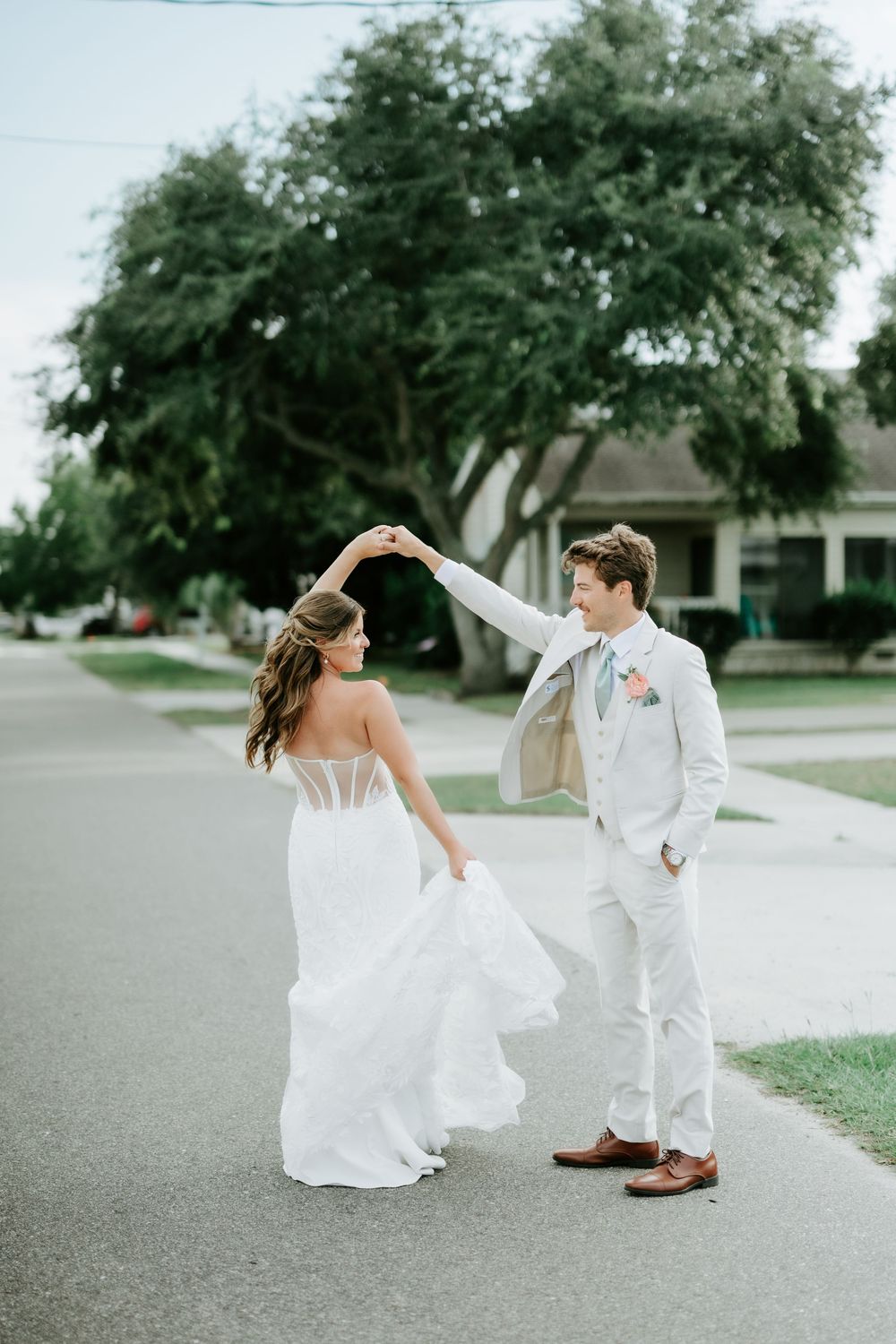 Bride in white gown and groom in light suit dancing on a suburban street with lush green trees in background.