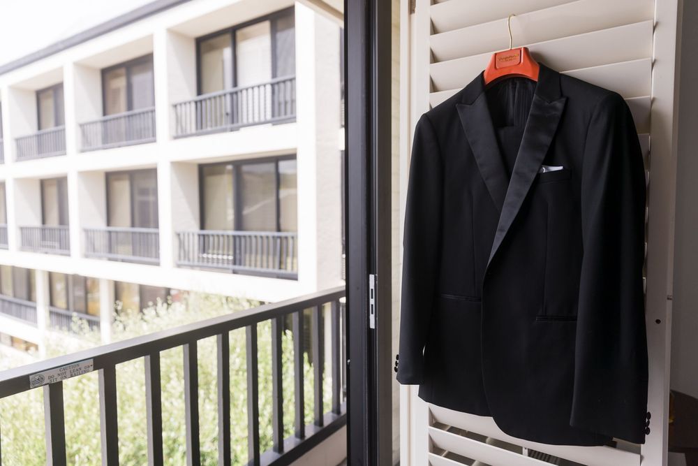 Black tuxedo jacket hanging on orange hanger over balcony door with hotel building in background.