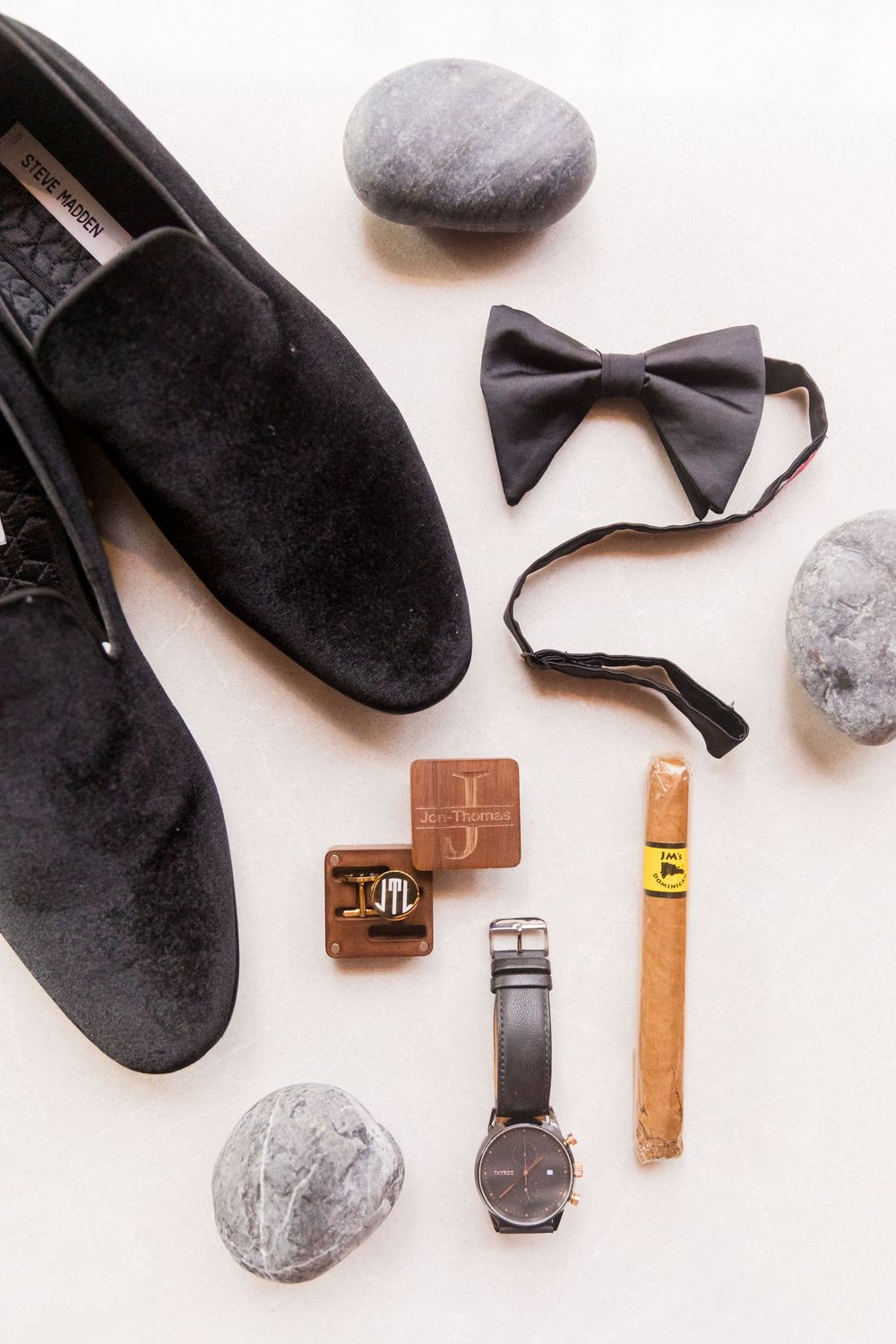 Grooms wedding flat lay featuring black velvet loafers, bow tie, watch, monogram cufflinks, and cigar on white background.