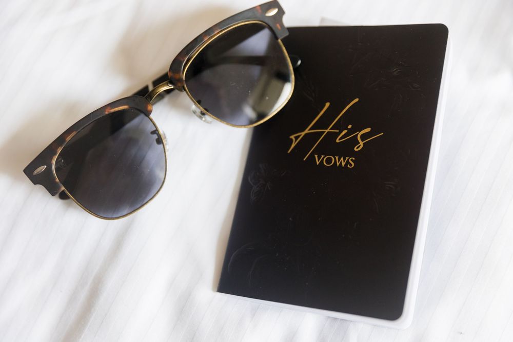 Black vow book labeled His Vows beside stylish tortoiseshell sunglasses on white background.