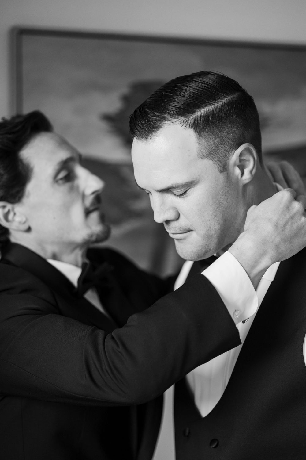 Black and white photo of a man adjusting the tuxedo collar of a groom during wedding preparations.