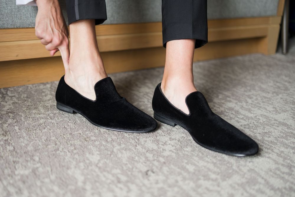 Man wearing black velvet loafers slip-on dress shoes, putting them on while standing on a gray carpet.