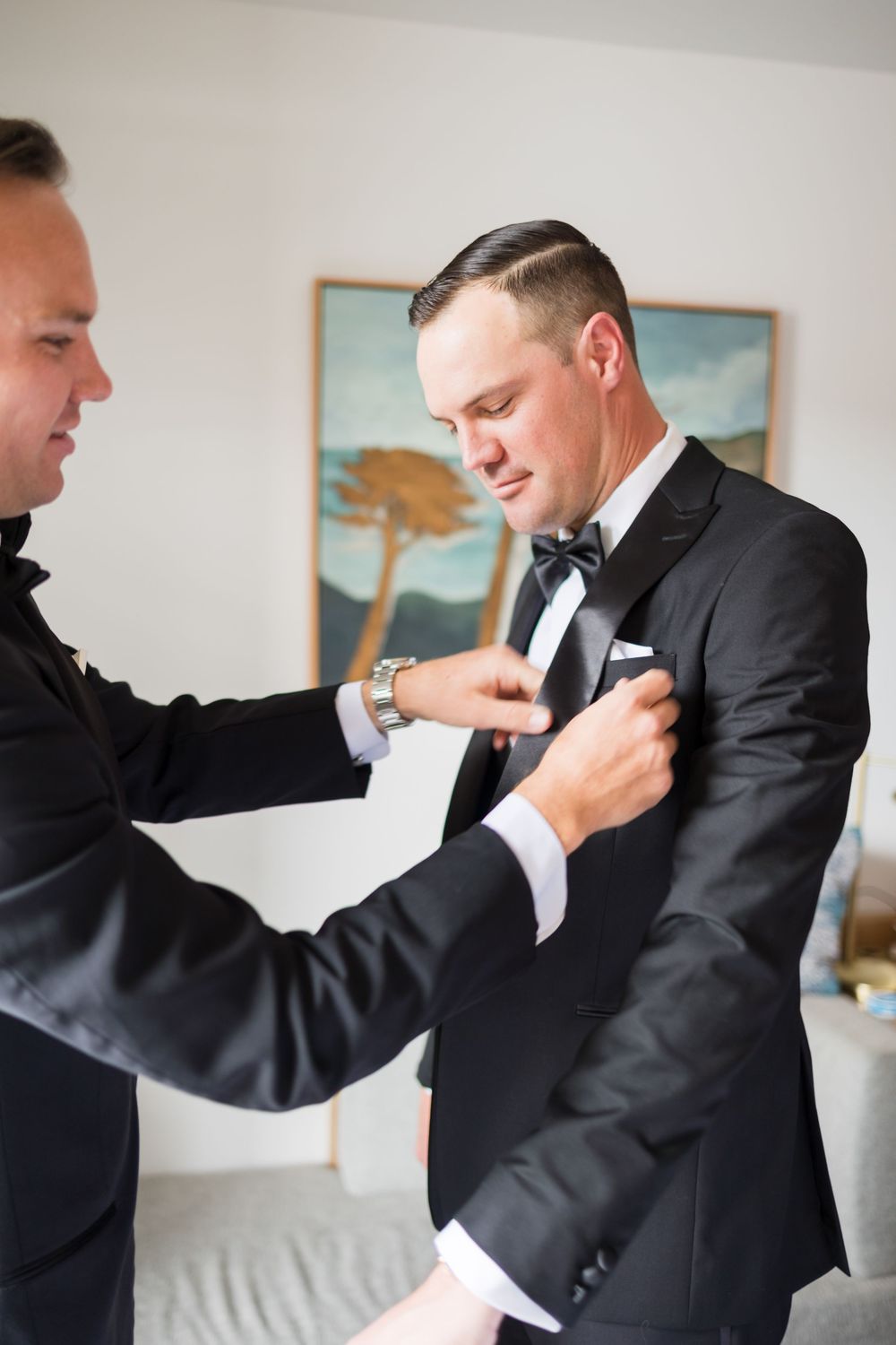 Groomsman helping groom adjust boutonniere on black tuxedo jacket before wedding ceremony.