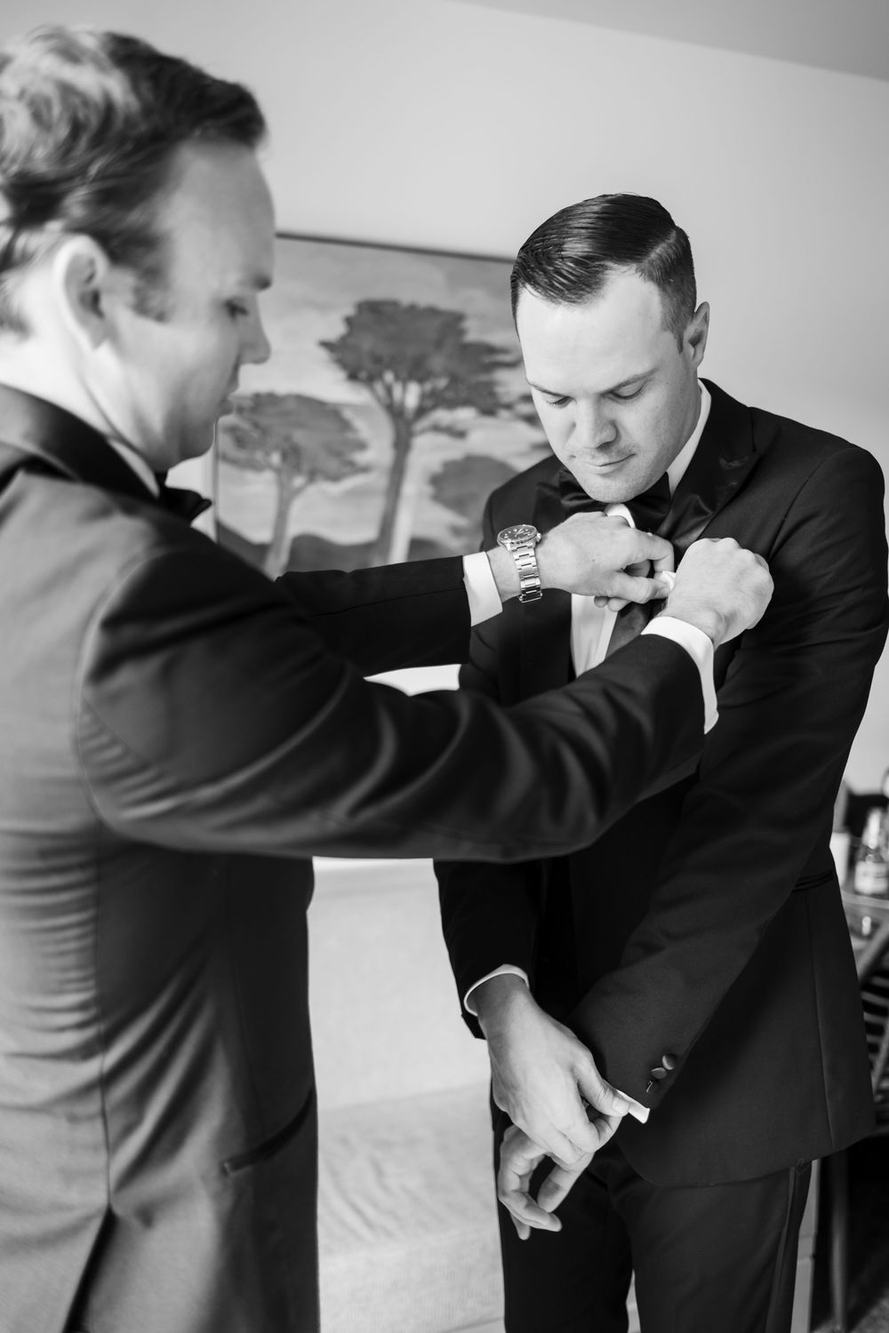 Groom getting his tie adjusted by best man while preparing for wedding ceremony in black and white photo.