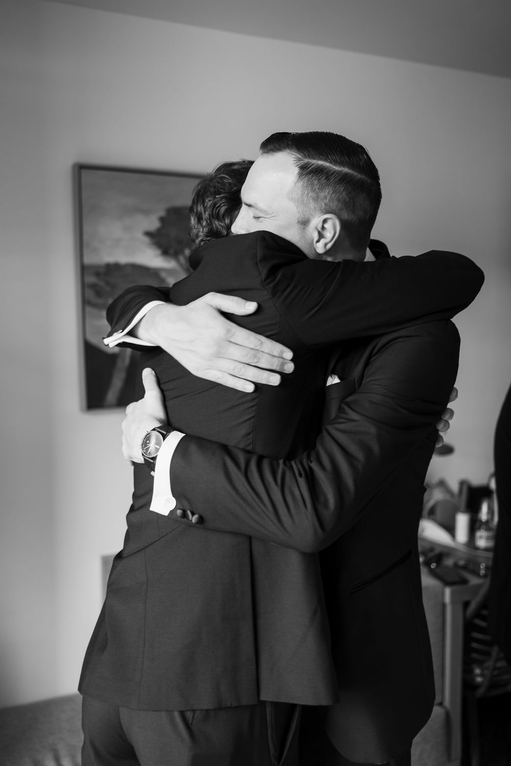 Two men in black tuxedos share an emotional embrace during wedding day preparations in black and white photo.