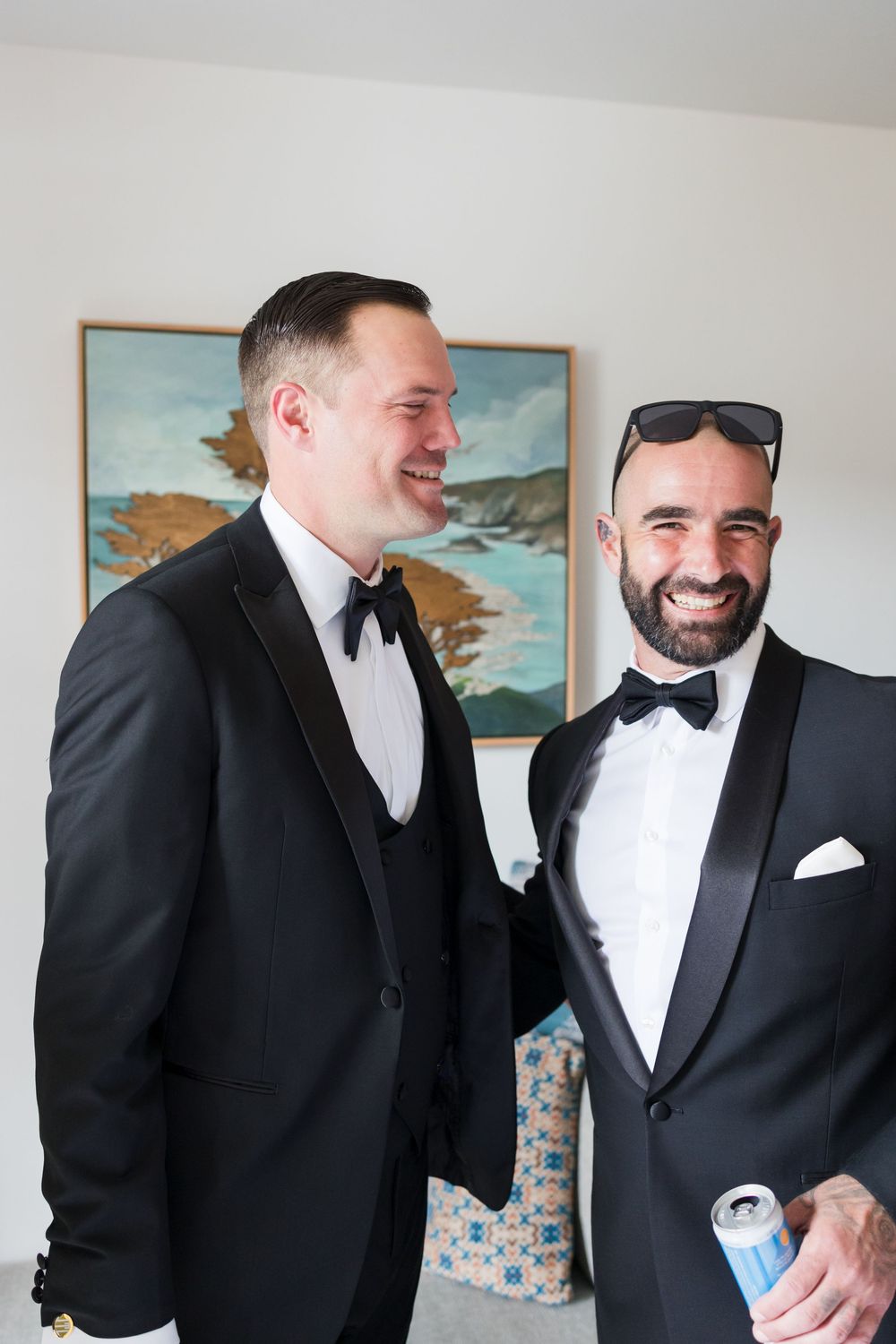 Two smiling groomsmen in black tuxedos with bow ties laugh together before a wedding ceremony.