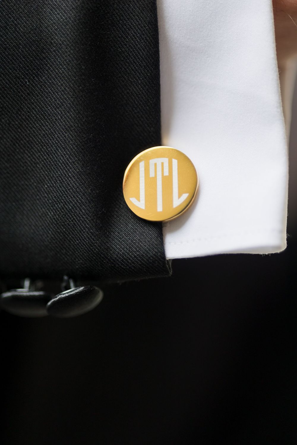 Gold monogram cufflink with initials JTL on white dress shirt sleeve beside black tuxedo jacket.