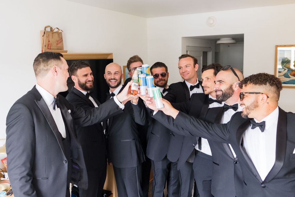 Groomsmen in black tuxedos toasting with whiskey bottle before wedding celebration.